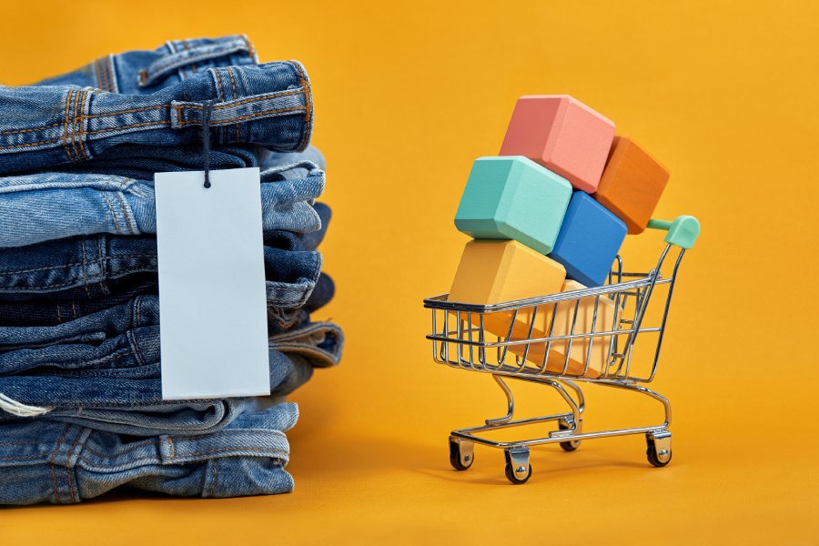 Carrinho de compras com pedidos e ao lado uma pilha de calças jeans, representando pedidos nos marketplaces que sofrem variação de preço.