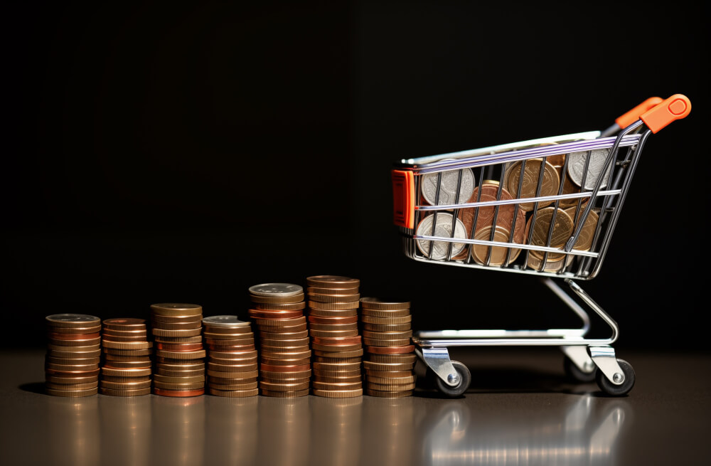 Carrinho de compras ao lado de pilhas de moedas, representando a precificação de produto e o impacto do preço na rentabilidade.