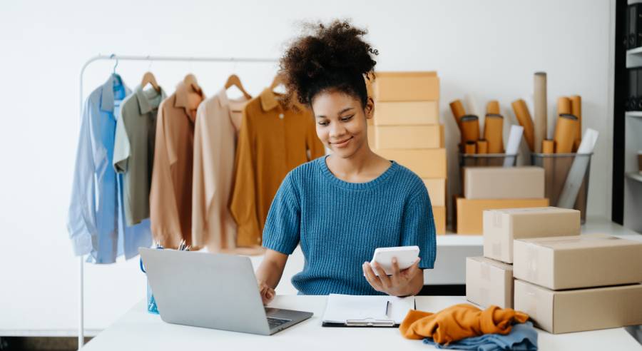 Vendedora de moda consultando celular e notebook em meio a caixas e peças de roupa, gerenciando precificação na Amazon