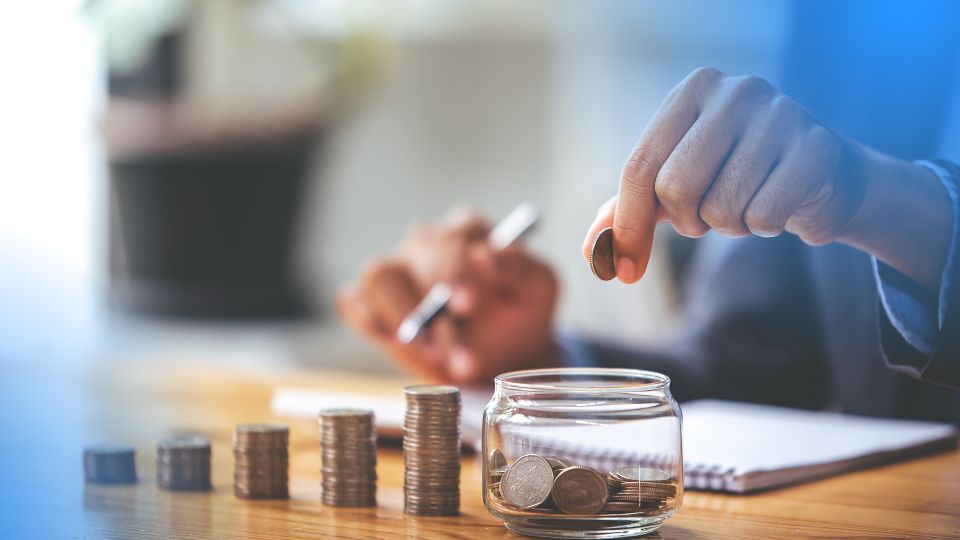 Mãos depositando moedas em pote de vidro com pilhas crescentes, ilustrando o cálculo de margem de contribuição por produto.
